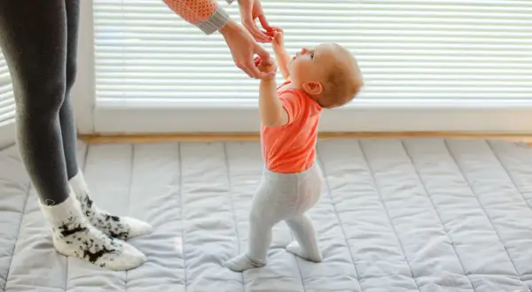 Mama pomaga dziecku, kiedy zaczyna chodzić. Maluch w pomarańczowej bluzeczce i szarych legginsach robi pierwsze kroki.