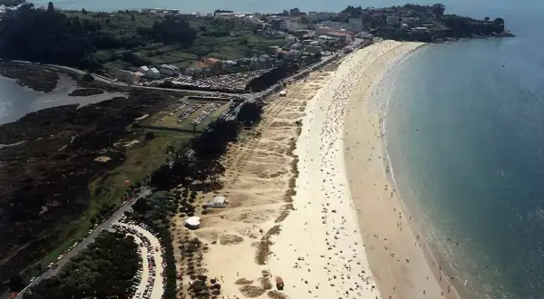 Vista aérea de una playa grande y concurrida, con arena dorada, mar azul y un pueblo costero al fondo.
