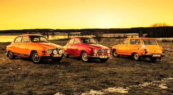 Tre klassiska Saab-bilar, en röd och två orange, står uppradade i solnedgången. En laban bil som väcker nostalgi.