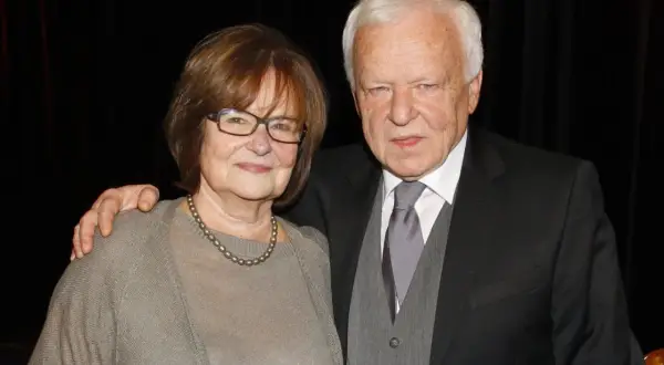 Anna Opani w okularach i z perłami na szyi, obok elegancko ubranego mężczyzny w garniturze.