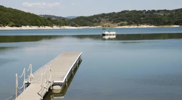 Un pontón blanco navega en el tranquilo embalse de Prada, con un muelle de madera en primer plano y colinas verdes al fondo.