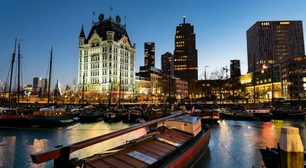 Wieczorem w porcie, gdzie jest Rotterdam. Widać zabytkowy budynek, nowoczesne wieżowce i zacumowane łodzie.