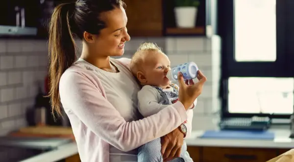 Uśmiechnięta mama karmi butelką niemowlę. Kiedy można podać koperek dla noworodka? To pytanie nurtuje wielu rodziców.