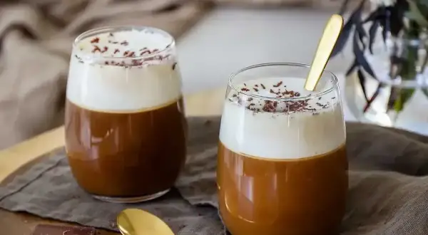 Två glas med krämig kaffe Karlsson, toppad med vispgrädde och chokladströssel. En gyllene sked ligger i ett av glasen.