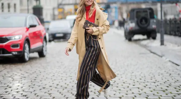 Frau in Beige-Trenchcoat, rotem Rollkragenpullover und gestreiften Hosen, die zeigt, wie man einen Trenchcoat kombinieren kann.