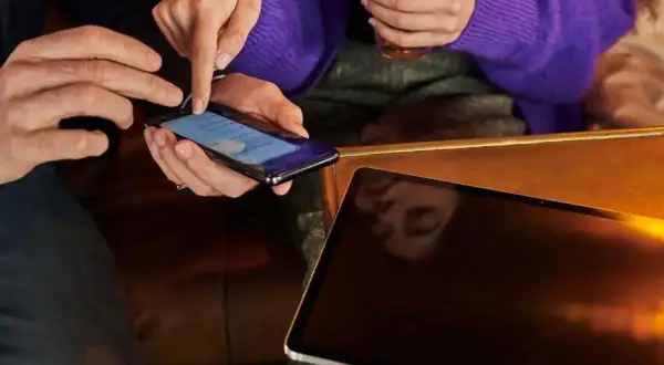 Dwie osoby przyglądają się smartfonowi, jedna z nich pokazuje, jak zwiększyć litery w telefonie. Obok leży tablet.