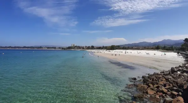 Amplia vista de la playa del Vao en Vigo, con aguas cristalinas, arena blanca y gente disfrutando del sol.