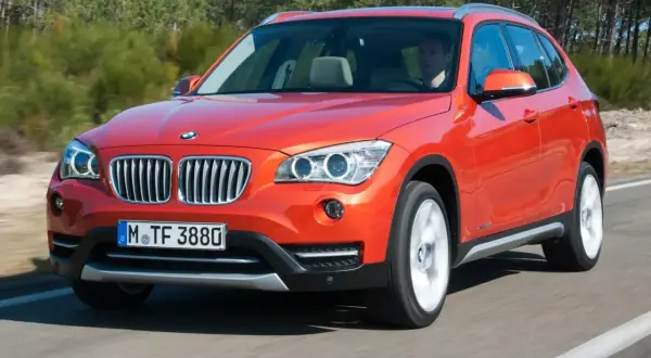 Pomarańczowy BMW X1 E84 w ruchu, z kierowcą za kierownicą, na tle drzew.