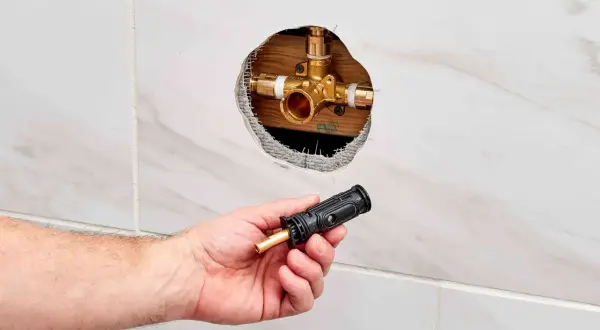 Hand holding a shower valve cartridge next to an open wall cavity revealing the shower valve body.