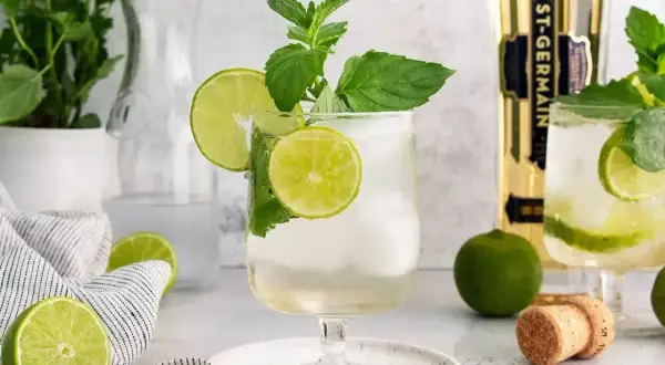 Orzeźwiający Hugo Spritz z limonką i miętą, idealny na letnie popołudnie. Obok butelka St-Germain.