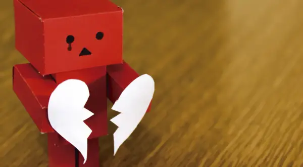 Un robot rojo de origami, con cara triste y una lágrima, sostiene las dos mitades de un corazón blanco. Parece estar a punto de morir de amor.