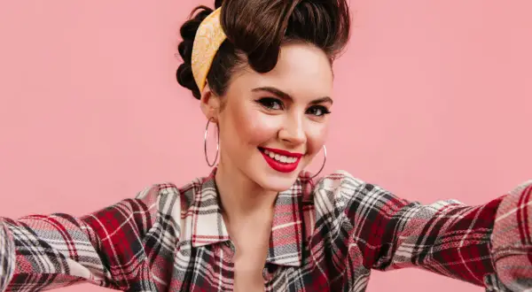 Uśmiechnięta kobieta w stylu pin up, z burzą brązowych włosów upiętych w kok i żółtą apaszką, w kraciastą koszulę i z dużymi kolczykami.