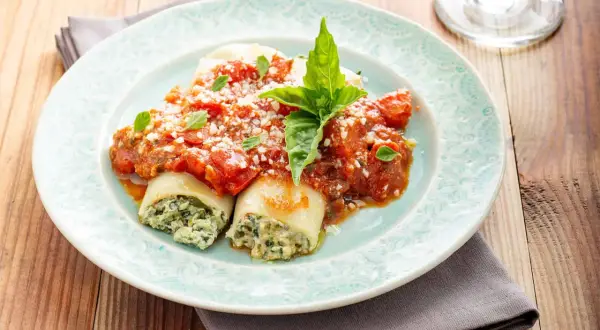 Canelones de espinacas cubiertos con salsa de tomate y queso rallado, adornados con hojas de albahaca fresca.