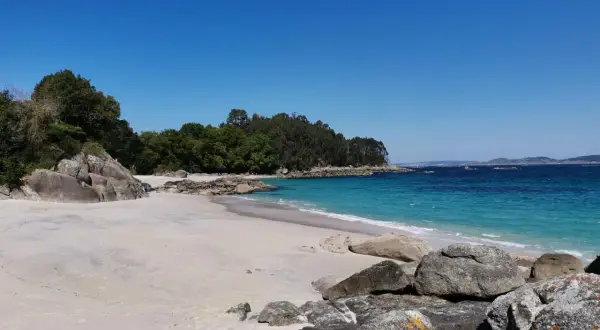 Arena blanca, aguas turquesas y rocas salpican la **playa de Castiñeiras**, un rincón idílico bajo un cielo azul intenso.
