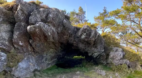Entrada a la cova da serpe, una cueva natural entre rocas y vegetación bajo un cielo azul.