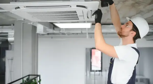 Instalator w kasku montuje klimatyzator kasetonowy na suficie.