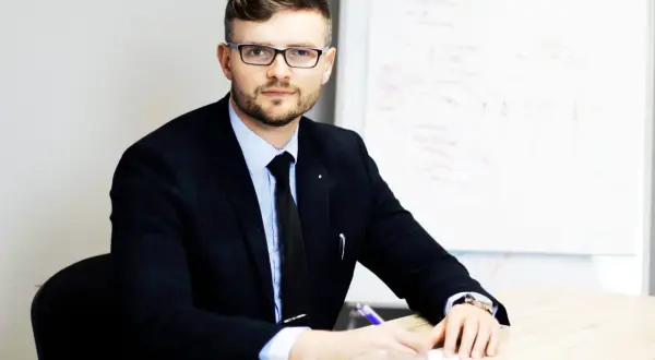 Mężczyzna w garniturze i okularach siedzi przy biurku, pisząc coś na kartce. Obok filiżanka kawy.