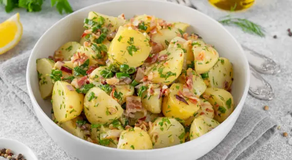 Pyszny kartoffelsalat przepis: ziemniaki z boczkiem, cebulką i ziołami w białej misce, obok cytryna i oliwa.
