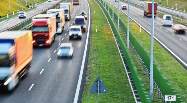 Autostrada z ruchem samochodowym i ciężarowym, zielone ekrany akustyczne i znak drogowy.