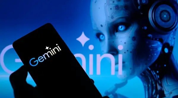 Ręka trzyma smartfon z logo Gemini. W tle widać twarz robota, która przypomina miękkie dildo.