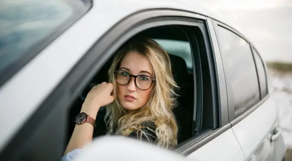 Kobieta w okularach za kierownicą białego samochodu. Zastanawia się, jakie okulary do jazdy samochodem wybrać, by zapewnić sobie komfort i bezpieczeństwo.