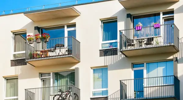 Nowoczesny budynek z balkonami, na jednym rower, na drugim stolik z krzesłami. W tle błękitne niebo, a na fasadzie cień rzucany przez balustrady, jakby prowadził do jezdni.