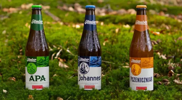 Trzy butelki piwa Amber: APA, Johannes i Pszeniczniak, stoją na mchu. Złote lwy z logo Amber są widoczne na butelkach.