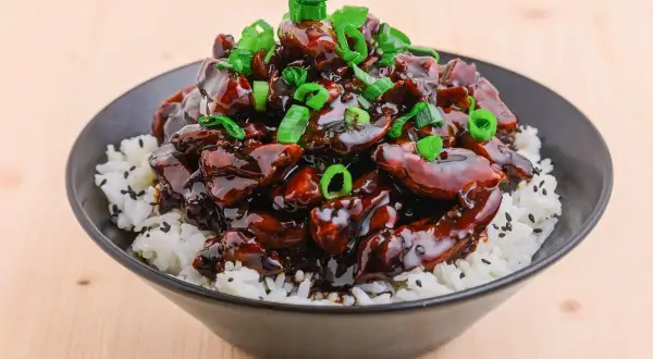 En skål med ris toppad med glansig teriyaki kyckling och strimlad salladslök.