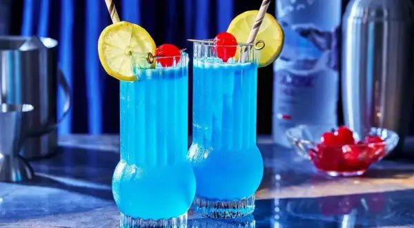 Dwa orzeźwiające drinki Błękitna Laguna, ozdobione cytryną i wiśnią, stoją na barze obok butelki wódki i shakera.