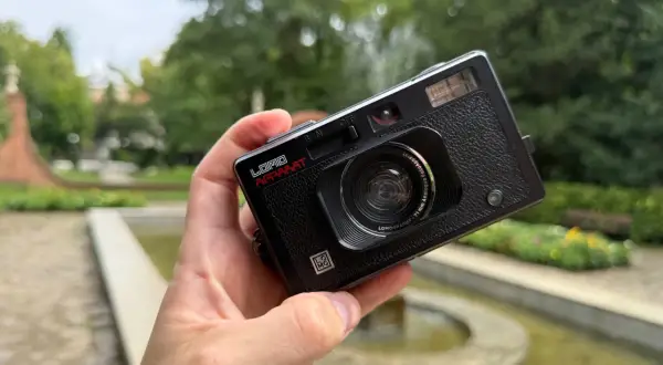 Czarna, analogowa lomografia LomoApparat z obiektywem 21mm. W dłoni, na tle zieleni i fontanny.