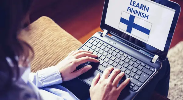Osoba uczy się języka fińskiego na laptopie, widoczna flaga Finlandii i napis "LEARN FINNISH".