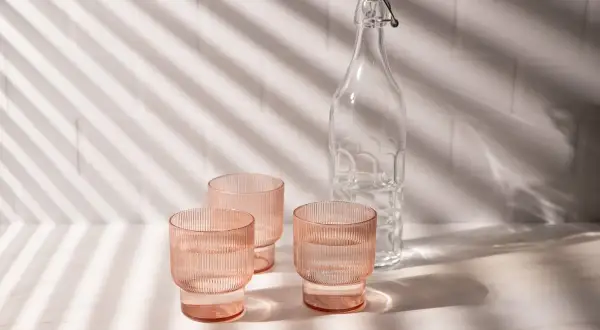 Natural product photography: three pink ribbed glasses filled with water and a clear glass bottle with a swing top, bathed in dappled light.