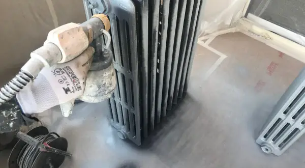 Proceso de pintar radiadores de hierro con pistola. Guante protege mano mientras se aplica pintura gris.
