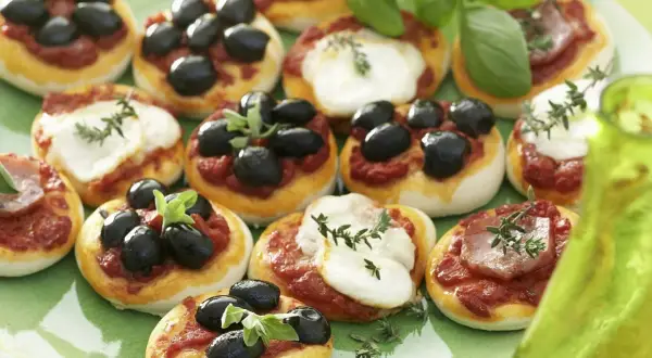 Kleine Pizzen mit Oliven und Mozzarella auf einem grünen Teller, garniert mit Basilikum.