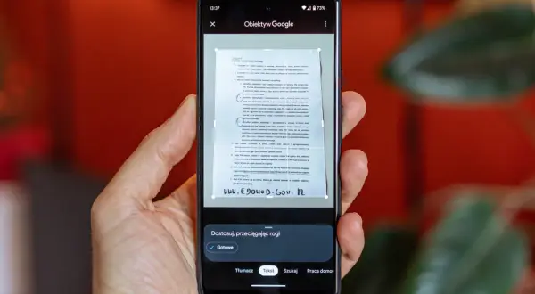 Ręka trzyma smartfon, na ekranie którego działa skaner dokumentów. Aplikacja Obiektyw Google skanuje tekst z papierowego dokumentu.