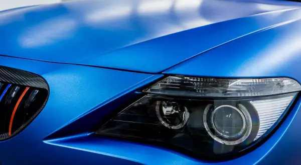 Matowy, **laguna blue** lakier na masce i błotniku sportowego auta. Widoczny fragment reflektora i grill z akcentami M.