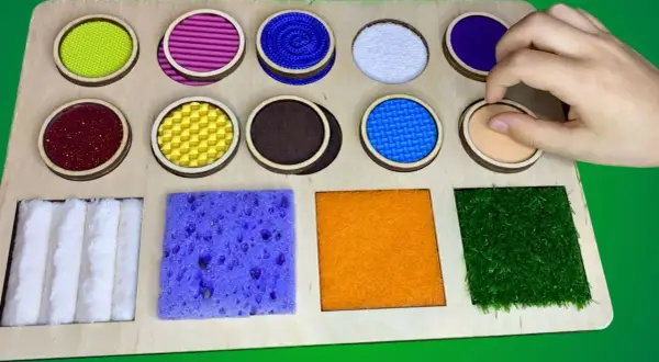 Tabla sensorial con diversos tipos de texturas para niños: círculos con materiales variados y cuadrados con esponja, fieltro y césped artificial.