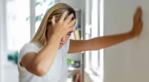 Femme blonde se tenant le front, appuyée contre un mur, le visage pâle, signe d'un malaise vagal.