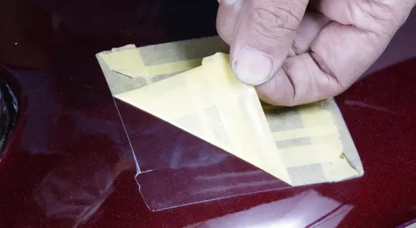 Una mano rimuove nastro adesivo da una superficie lucida color bordeaux. Il trasparente sulla vernice serve a proteggerla da graffi e usura, mantenendone la brillantezza.