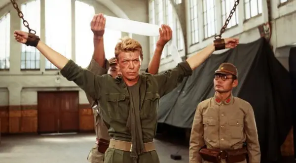 David Bowie jako major Celliers, pojmany w "Merry Christmas Mr. Lawrence", z rękami przykutymi do łańcuchów. Obok stoi japoński oficer.