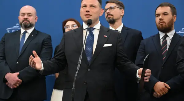 Mężczyzna przemawia przy mikrofonie, otoczony innymi politykami. W tle niebieska ściana z napisem. Konfederacja postulaty.