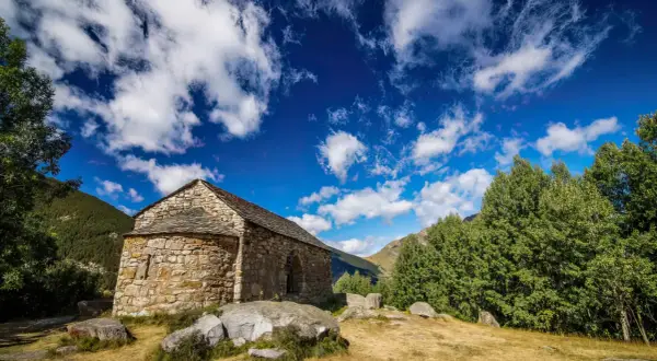 Pequeña ermita de piedra en las rutas del Vall de Boí, rodeada de naturaleza y bajo un cielo azul con nubes algodonosas.