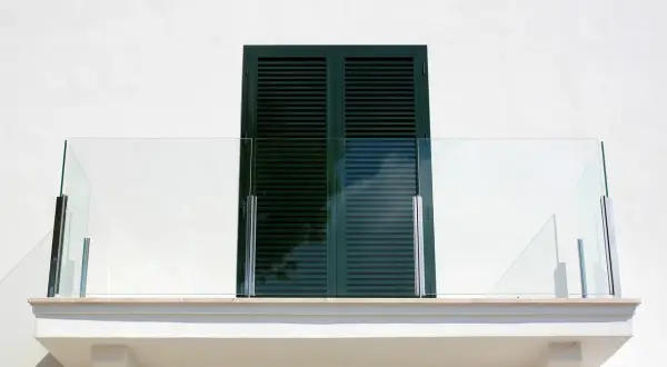 Nowoczesny balkon z przeszkloną balustradą i ciemnozielonymi okiennicami. Okapnik balkonu zapewnia ochronę przed deszczem.