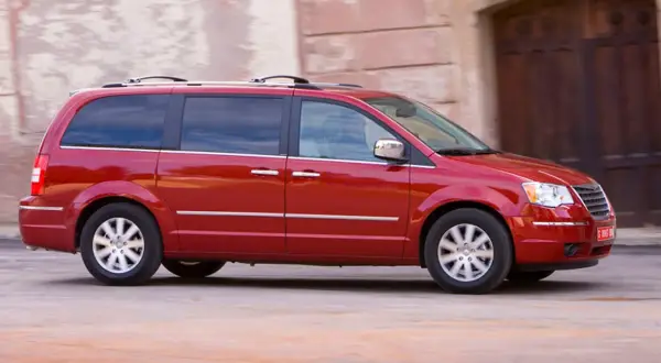Czerwony Chrysler Grand Voyager w ruchu, z przyciemnianymi szybami i relingami dachowymi, przemierza ulicę.