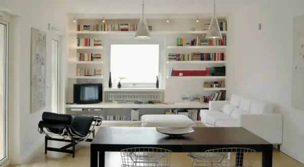Soggiorno moderno con ampie librerie cartongesso a parete, piene di libri, che incorniciano una finestra. Divano bianco e poltrona nera completano l'arredo.