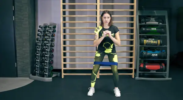 Kobieta w sportowym stroju wykonuje ćwiczenie monster walk z gumą oporową na udach.