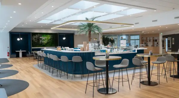 Un espace restauration moderne avec un bar bleu, des tabourets gris, une plante tropicale et des tables rondes.