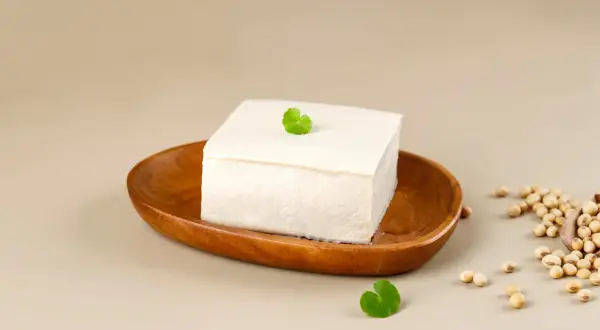 Kostka białego tofu na drewnianej misce, obok nasiona soi. Tofu powstaje z ziaren soi.