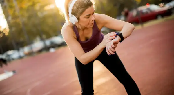Kobieta w słuchawkach sprawdza czas na zegarku sportowym, przygotowując się do biegu na 10 km. Czas amatora jest ważny!