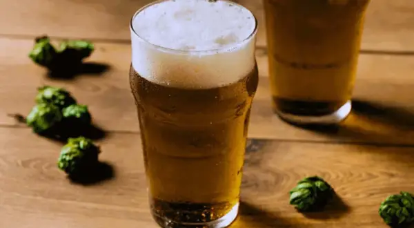 Un bicchiere di birra dorata con schiuma abbondante, circondato da luppoli verdi su un tavolo di legno. Perfetto per una festa della birra!